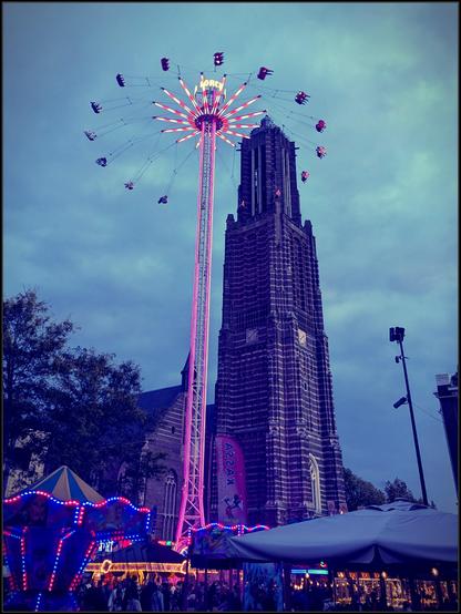 A very high church tower looming against a darkening and overcast sky. In front of it is a colorful and brightly lit kind of chairoplane, which is lifted so high into the sky that it looks to be nearly as tall as the church steeple.