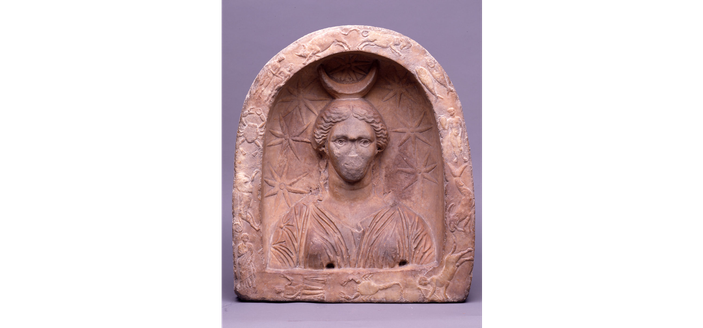 Reddish marble carved votive relief of Selene: a female bust to the front in an arched niche, draped in a sleeved chiton, and with her hair parted in the middle and brought down over her ears, nose now missing, surrounded by a crescent on her head, seven stars in the field around and the signs of the zodiac in low relief; inscribed beneath with an unitelligible Gnostic formula and noted in the catalogue that from its position the inscription appears to have been an afterthought. 