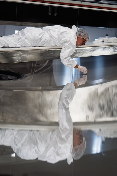 Most of this image is taken up by the silvery, reflective surface of an enormous mirror. An ESO staff member in all-white, transparent plastic hat covering their head and similar gloves on their hands. They reach out over the mirror surface, something small hidden in the palm of their hands. As they lightly touch the mirror surface, a slightly distorted reflection appears to reach back towards them, showing a near-perfect mirror image on its pristine metal.