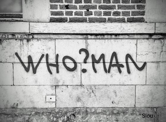 Photo noir et blanc.
Sur un mur dans la rue est inscrit à la bombe :" Who?man"