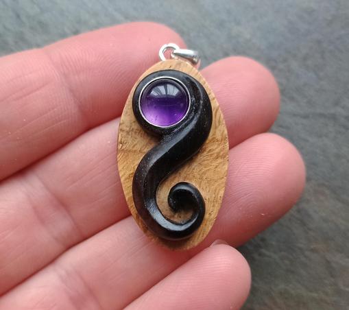 A round purple amethyst stone, set within a swirl of hand carved ebony. Mounted to an oval of golden brown burl oak. With a silver bail necklace attachment at the top. Resting on the fingertips of a band, with a grey slate background.