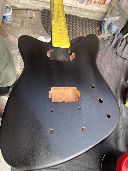 Eine schwarze E-Gitarre in Arbeit, von oben fotografiert, nur der Korpus und ein Stück des mit gelbem Klebeband abgeklebten Halses zu sehen.
A black electric guitar, view from above. Only the body and a part of the neck, taped with yellow maskingtape, are visible.