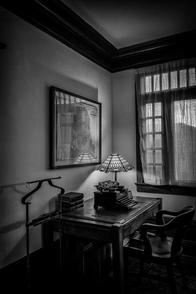 Corner of a apartment in San Francisco, furnished to look like it might have in the late 1920s. We see a wooden desk with an old typewriter, books and an travel clock on it. Above the desk is a map of San Francisco.