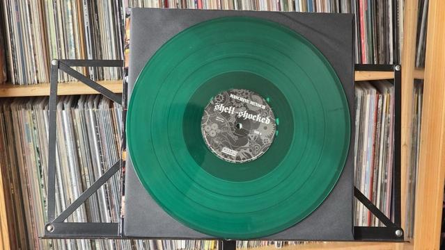 Eine grüne Schallplatte mit dem Titel "Shell Shocked" und einem schwarzen Etikett mit verschlungenen Mustern. Sie wird auf einem Ständer mit einem Hintergrund aus ordentlich angeordneten Schallplatten präsentiert.