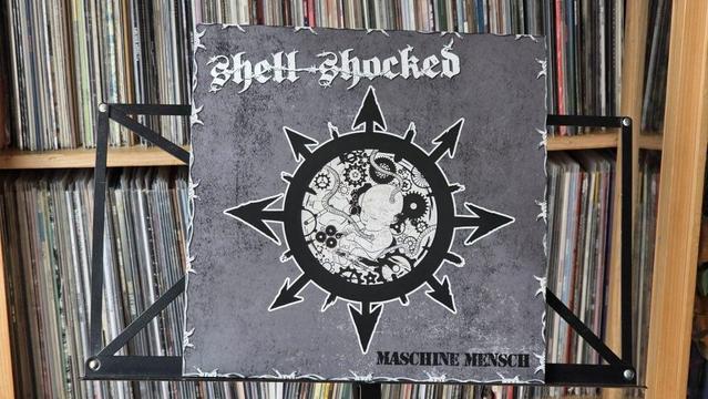 Das Bild zeigt das Albumcover mit dem Titel "Maschine Mensch" von Shell Shocked. Das Design umfasst eine zentrale Grafik, umgeben von Pfeilen auf einem strukturierten Hintergrund. Es ist auf einem Ständer vor einer Sammlung von Schallplatten abgebildet.