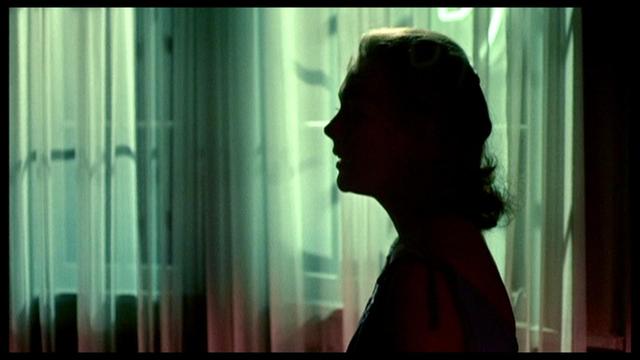 A silhouette of a woman against a backdrop of softly illuminated curtains, featuring a blend of green and red hues. The light casts a moody atmosphere, highlighting her profile.