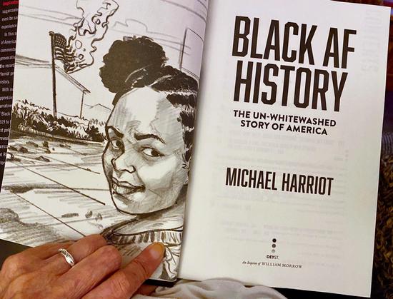 A photo of my hand holding open my copy of the book titled "Black AF History: The Un-Whitewashed Story of America" by Michael Harriot. The left page features an illustration of a woman smiling with a burning American flag in the background. This is the book I am currently reading.