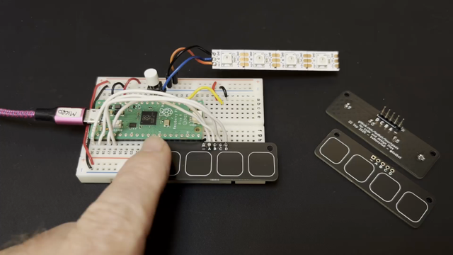 Solderless breadboard with a Raspberry Pi Pico, a four-LED WS2812 strip, and a black PCB with four capsense touch buttons. As a finger presses the buttons, the corresponding LED lights up