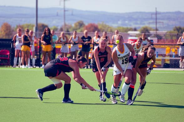 In the middle of an almost featureless green turf five girls struggle for control of a ball with candy-cane shaped sticks while more girls look on from the sideline, power lines and a distant line of hills can be seen in the far distance.