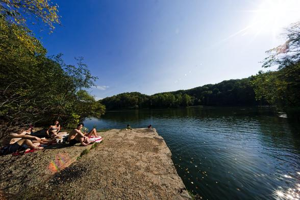 A group of asian students rest on a large outcropping of rock near a large pond with a forested distant size and sunstar reflections in the lower left corner.