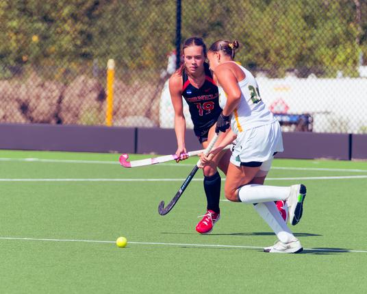 A girl with number 19 in red  on a black jersey with black socks and red shoes is facing us holding a field hockey stick while number 21 of the opposing team in green in a white uniform cuts in from the right,  behind there is a black chain link fence,  tall grass,  and further away trees