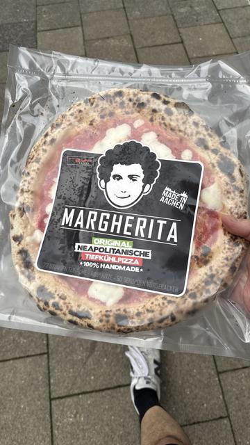Das Bild zeigt eine Pizza mit einer Beschriftung, die "MADE IN AACHEN" in großen, weißen Buchstaben auf einem schwarzen Hintergrund zeigt. Die Beschriftung ist von einer weißen Silhouette der Stadt Aachen umgeben, die die charakteristischen Türme und Dächer der Stadt darstellt. Die Pizza selbst ist sichtbar, mit rotem Tomatensoße und weißen Käseflocken, die auf dem Teig verteilt sind. Der Rand der Pizza ist leicht gebräunt, was auf eine knusprige Kruste hinweist. Die Beschriftung und die Pizza …
