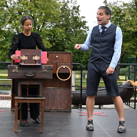 Sia und Thomas stehen auf einer Open Air Bühne. Vor ihnen ist ein improvisierter Tisch aus einem Stuhl, einem Hocker und einem alten Koffer aufgebaut. Auf dem Koffer liegt ein geöffnetes Pop-Up Buch. Es zeigt zwei Hände in Klatschposition, die durch ein Ziehen an der linken Buchseite auch richtig klatschen.