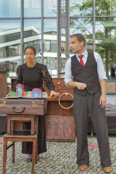 Sia und Thomas stehen wieder neben bzw. hinter dem improvisierten Tisch. Vor ihnen das geöffnete Pop-Up Buch. Aus dem Buch ist eine Jahrmarktsszene mit Riesenrad und Schaubude gewachsen.