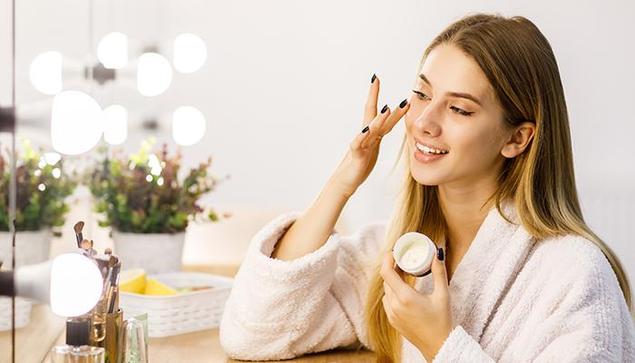 A young woman in a bathrobe applying face cream in front of a mirror as part of her Korean Skincare Routine for glowing and healthy skin.