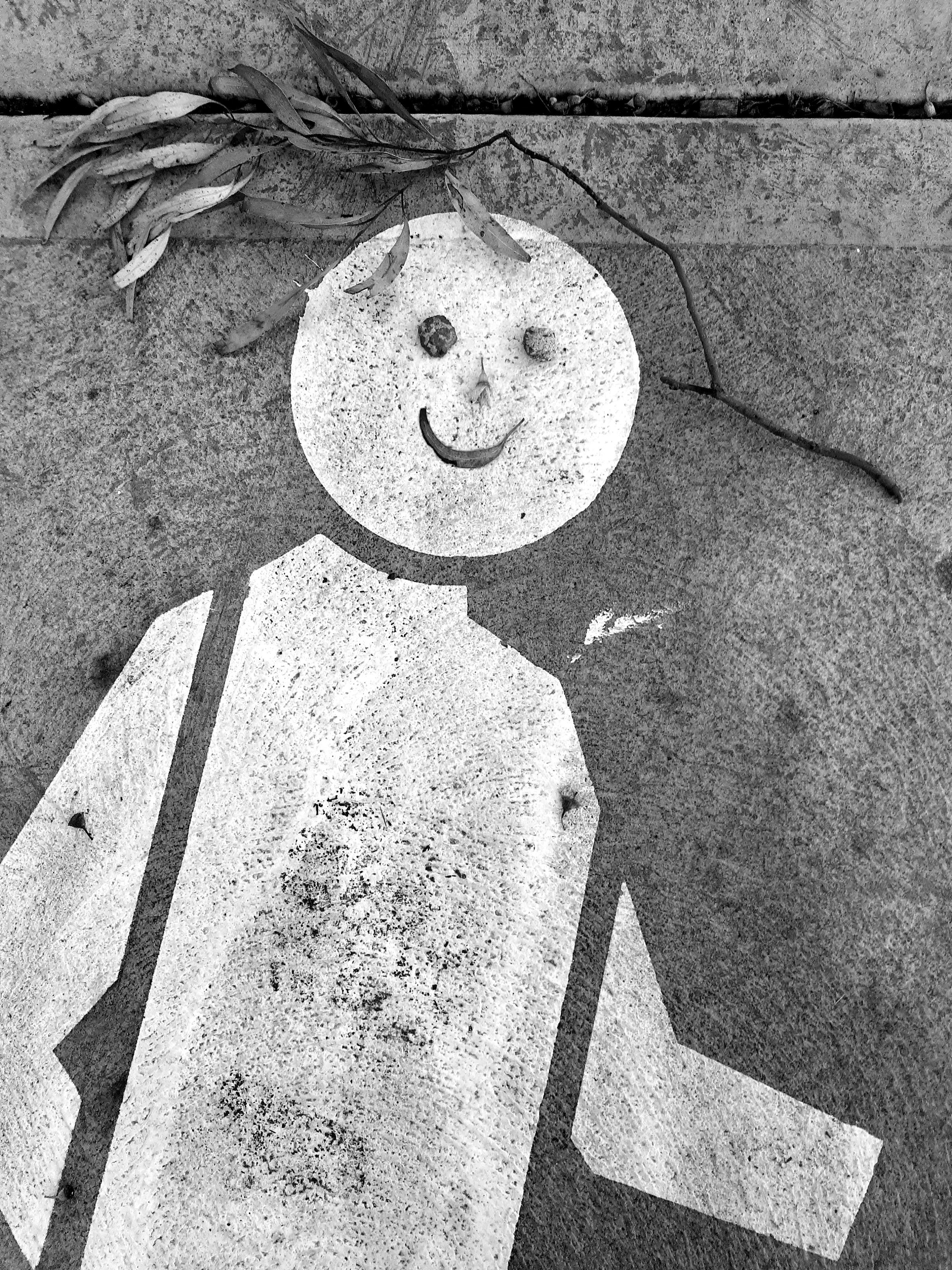 A stencilled sidewalk sign in a "walking" pose. We see it from above, white upon a grey pavement.  Inside the solid circle of the head, someone has placed two pebbles, a tree seed, and a curled leaf, to signify a smiling face. Above the head is a small branch of leaves, resembling hair