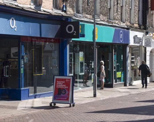 Britsh street with three self-phone shops in a row:  O2, BT and Three.  O2 has a magenta sign in front of it,  a woman is standing near the entrrance to the BT store and a man seems to be walking past Three.