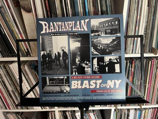 Das Bild zeigt das Cover einer Vinylplatte mit dem Titel "Blast Off NY" von Rantanplan, die auf einem Notenständer steht. Das Design enthält eine Mischung aus Fotos, darunter Bandmitglieder und Studioszenen, mit leuchtenden Farben und Text, der den Titel des Albums hervorhebt.