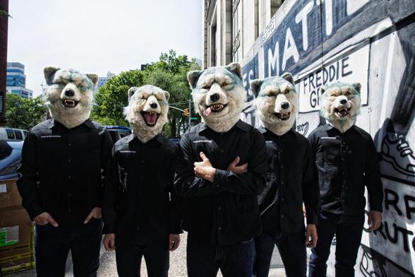 The photo shows the five members of MAN WITH A MISSION standing in a row on a street. They are dressed in dark clothing, including jeans and shirts with a MAN WITH A MISSION on it, while wearing their full-face wolf masks.