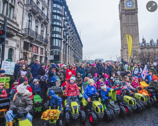 Protest on the streets in the UK with people standing in the back and people riding mini-tractors in the front.