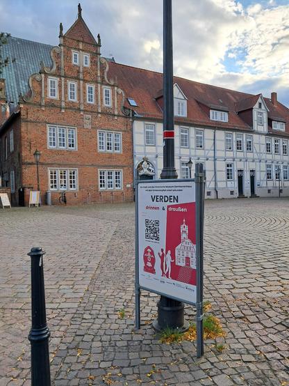 Ein Platz mit historischen Gebäuden (teils Fachwerk), im Vordergrund ein Laternenpfahl mit einem Aufsteller in weiß-pink, auf dem Groß "Verden drinnen & draußen" steht
