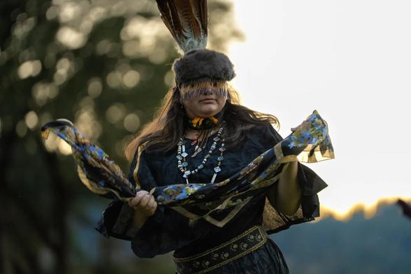 A person in traditional regalia with feathers, beadwork, and a patterned shawl performs a dance outdoors at sunset.