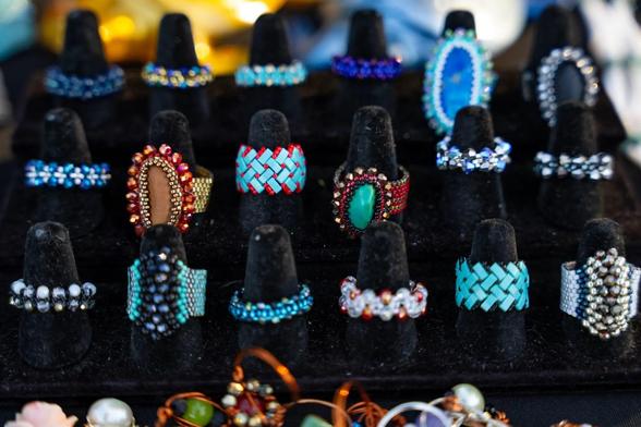 A display of colorful handmade beaded rings, featuring intricate patterns and gemstones, arranged on black ring stands.