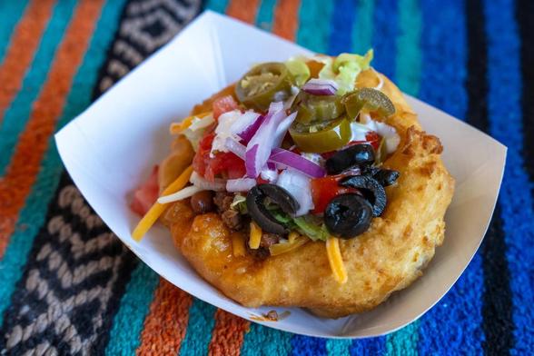 A loaded fry bread taco topped with cheese, lettuce, onions, tomatoes, olives, and jalapeños sits in a paper tray on a colorful woven blanket.