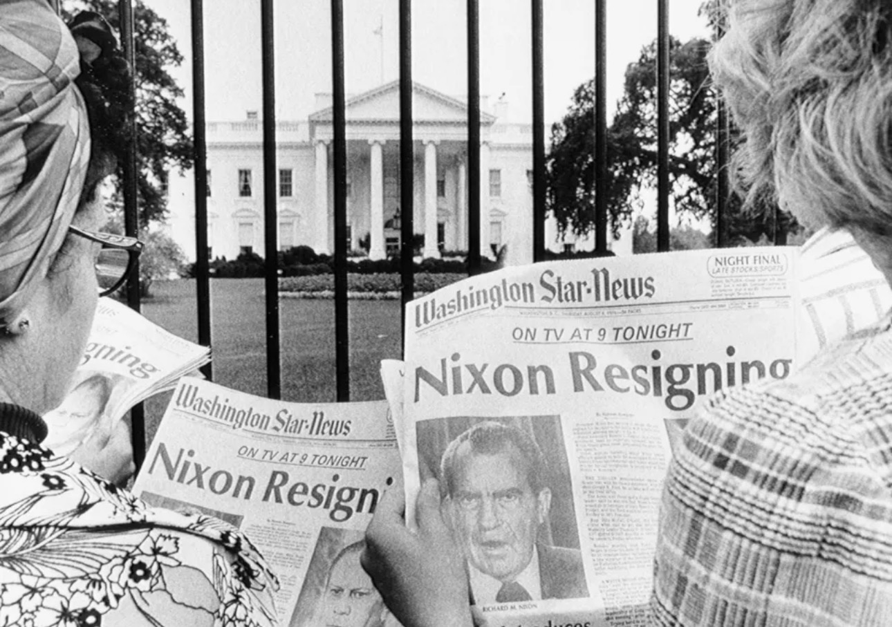 Black-and-white photograph of two people reading newspapers outside the White House gates in August 1974. The front pages of the Washington Star-News headline “Nixon Resigning” with a large photo of President Richard Nixon. The White House is visible in the background beyond the iron fence.