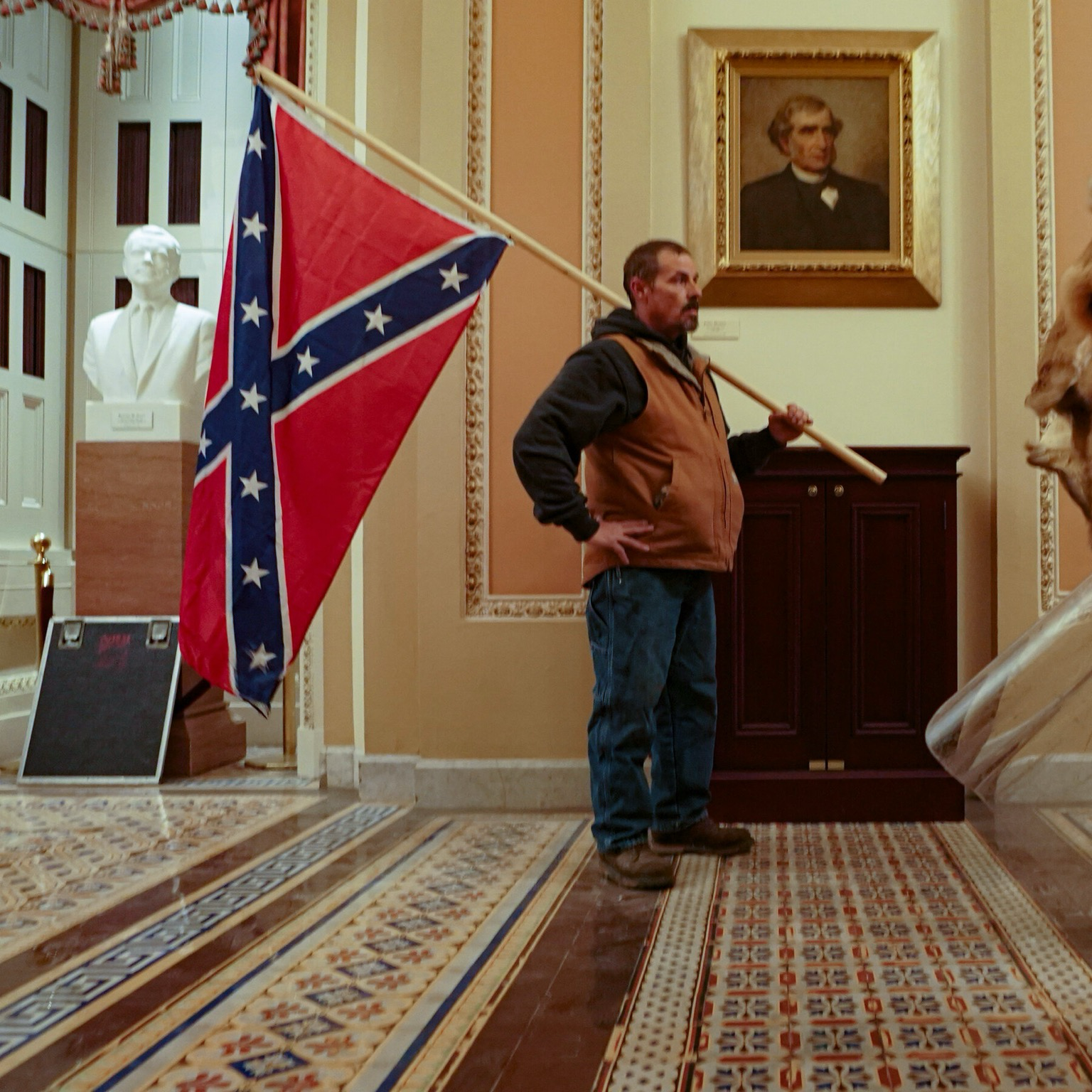 A man stands inside the U.S. Capitol holding a large Confederate flag on a pole during the January 6, 2021 insurrection. He wears jeans, boots, a brown vest, and a black hoodie. Behind him are statues, ornate tile floors, and a portrait of John C. Calhoun hanging on the wall.