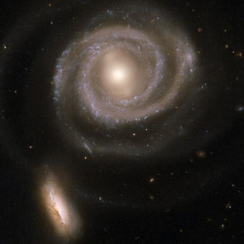 A pair of galaxies, a large face-on barred spiral galaxy and a smaller edge-on disk galaxy. The upper portion of the frame is dominated by the spiral galaxy NGC 5754. It has a bright, pale yellow, circular core that transitions into an almost vertical bar structure. Two well-defined spiral arms circle around the core counter-clockwise, beginning at the ends of the bar. The arms both complete one revolution around the core and then fade out. They are dotted with pale blue star clusters. At about 5 and 7 o'clock on the bottom of the galaxy, the spiral arms have small kinks, creating what looks like straight segments in the otherwise smoothly curving arms. Overall, the galaxy looks a little like a cinnamon roll. In the bottom left corner of the frame is the smaller companion galaxy NGC 5752. It is oval-shaped, with the long axis almost vertical, running parallel to the bar of the large spiral. On a background of pale yellow stars is a line of blue dots, which are star clusters, running through the long axis in the center of the galaxy. Below this is a dark brown dust lane. 