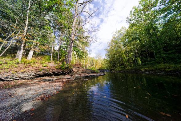 I'm standing in the middle of a shallow spot in the creek with ripples radiating away from my (just barely) unseen legs,  to the left is a close outcropping of rock with a flat part that then rises up and gets covered with trees and on the other seen a thin layer of rock is visible and lots of trees,  in the center of the frame the path curves out of sight first to the right and then to the left and the sky is mostly white cloud with just a little blue visible.