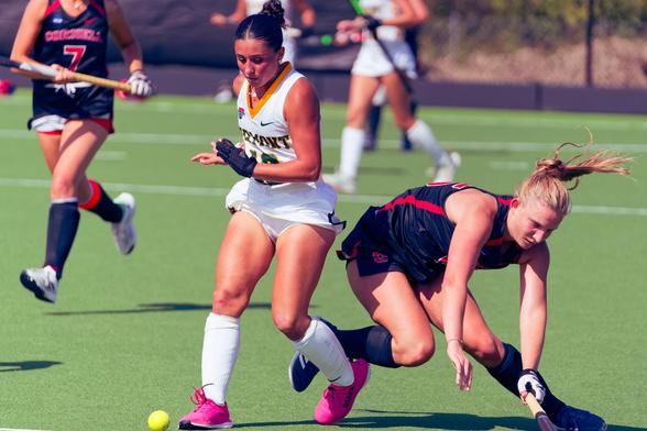 Two field hockey players seem to have collided at the legs,  both running in my direction.  One wearing a white jersey, white sockets and purple shoes is standing with the ball right in front of her toe,  the other wearing a black jersey with red trim is heading for the ground,  other players,  partially visible, seem to be converging on the scene