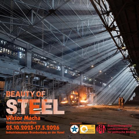 Siemens-Martin-Ofen in Brjansk. Ein Siemens-Martin-Ofen mit drei Türen in einer dunklen Industriehalle. Aus den Türen schlagen Flamme. Durch Öffnungen in der Decke leuchten Sonnenstrahlen. Am rechten Bildrand stehen zwei Arbeiter.