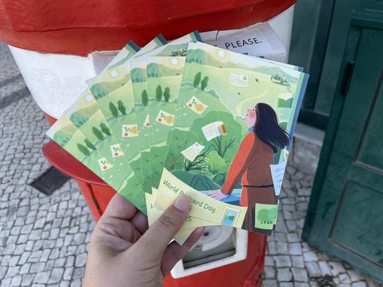 A hand holds several postcards in front of a bright red postbox. The postcards feature the official 2025 World Postcard Day design, showing an illustration of a woman outdoors, looking up as postcards float through a green landscape with trees and hills.
