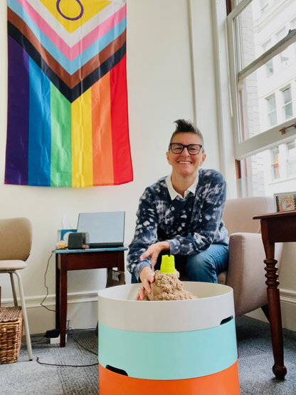 Image ID: a white gender queer lesbian, with short hair and dark rimed glasses smiles. They are wearing a blue and white sweatshirt. They have one hand on the kinetic sand and they sit in front of a 2SLBGTQIA+ progress flag.