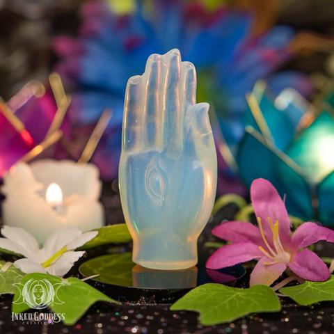 An example of the "Opalite Carved Hand with Eye" product stands upright on an obsidian disc. This opalescent glass is carved into the shape of an open hand with a vertical eye in the palm. Presented by Inked Goddess Creations.