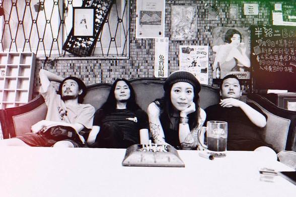 The photo shows the four members of Hibushibire casually relaxing on a sofa with a table in front of them. The photo in black-and-white with a colourshift filter showing a bit of red and green. While the three men are leaning back on the sofa, the female band member is leaning forward. On the table are an ashtray full of cigarettes and a drink.