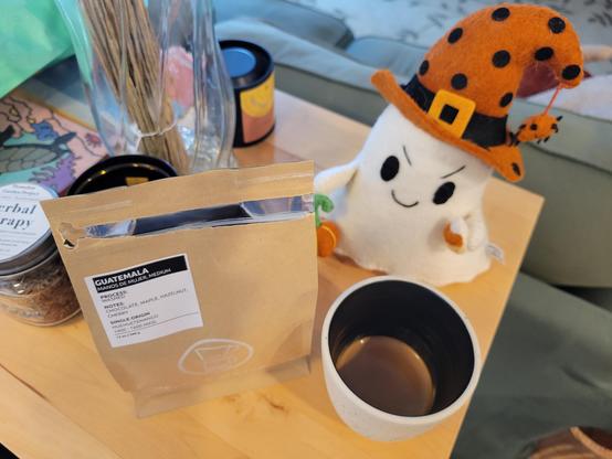 12-ounce, handle-less, off-white ceramic cup with a espresso shot, on a birch console table behind a green couch. The clay of the cup has many tiny flecks of black and blue. The interior of the cup has a dark-colored glaze.

To the left of the cup is a 12-ounce brown coffee pouch. It has a rectangular medium-sized white sticker at upper-left with coffee details. A white circular logo at bottom-center shows a pour-over filter holder over a transparent coffee mug. Text at bottom reads "Coffee & Water Lab" and "Single Origin - Roasted In Store" (white sans-serif block letters).

The text of the white sticker reads:
Header: "Guatemala, Manos De Mujer, Medium" (white sans-serif block letters on black background)
Process: Washed
Notes: Chocolate, Maple, Hazelnut, Cherry
Single Origin: Huehuetanango 1400-1600 MASL
12 oz/340 g

Other objects on the birch coffee table: a smiling 12-inch felted ghost with angled eyebrows which give it a fierce-but-friendly expression, with a witches hat (orange with black polka-dots) upon which a smiling spider dangles, holding a pumpkin and a candy corn. Also a glass face with lavender stems, a jar with aromatic herbs, a black metal candle holder, and a candle in a closed metal container. 