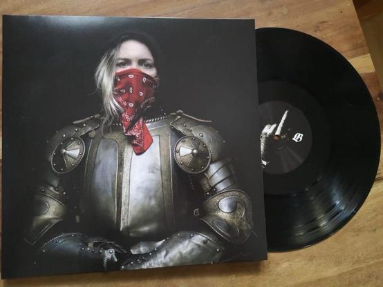 Ein Albumcover, das eine Person in einer mittelalterlichen Rüstung zeigt, die ein rotes Kopftuch um das Gesicht trägt. Neben dem Cover liegt eine schwarze Schallplatte auf einer Holzfläche.