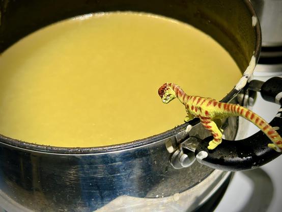 A green Dilophosaurus with brown spots stands on the handle of a large pot and gazes at the inside filled with yellow, congealing grease that has been treated with a hardening additive for easy disposal.