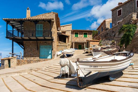Das warme, sonnige Gefühl eines kleinen Fischerdorfs in Spanien. Man fühlt sich sofort in einen Urlaubstag auf der spanischen Insel Mallorca versetzt, der Geruch von Salz in der Luft und das Zwitschern der Möwen sind fast greifbar.
​Das Foto zeigt eine felsige Bucht, in der einige Fischerboote an Land gezogen wurden. Das auffälligste Boot im Vordergrund, mit dem Namen "EN GROS" auf der Seite, liegt auf einer betonierten Rampe, die zum Wasser führt. Neben ihm liegen noch ein paar andere Boote, manche sind mit Planen abgedeckt. Im Hintergrund schmiegen sich alte Steinhäuser an den Hang, mit typisch spanischen Details wie blauen und grünen Fensterläden. Die Steinfassaden der Gebäude leuchten warm in der Sonne. Eines der Häuser hat einen Balkon mit einem Geländer aus dunklem Metall.