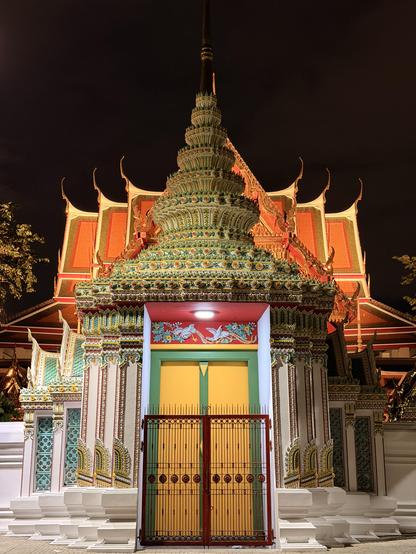 Verschlossener Eingang zum Tempel Wat Pho bei Nacht. Ein fein ziseliertes rotes Gitter versperrt die geschlossene gelbe Tür mit grünem Rahmen. Sie würde durch den weißen Eingang mit mehreren Säulen und tollem Spitzdach aus buntem Porzellan zum Tempel führen, dessen orangene Giebeldächer gerade noch zu sehen sind