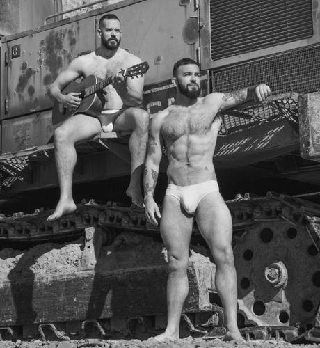 Two muscular men with beards who wear only white briefs are posing in front of a bulldozer. One of them is playing a guitar