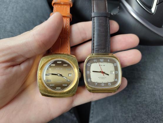 Ruka držící dvoje hodinky PRIM. Vlevo s hranatým zlatým pouzdrem a béžovo hnědým ciferníkem s manuálním nátahem. Vpravo PRIM Televize se zlatým hranatým pouzdrem a zaobleným čtvercovým ciferníkem s šedým okrajem s číslicemi a krémovým středem.