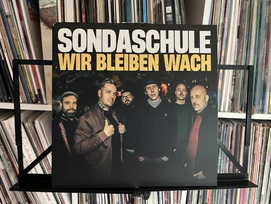 Das Albumcover für "Wir bleiben wach" von Sondaschule zeigt eine Gruppe von sechs Männern, die zusammen posieren. Im Hintergrund ist eine Sammlung von Schallplatten zu sehen. Der Titel des Albums ist in großen, fetten Buchstaben prominent dargestellt.
