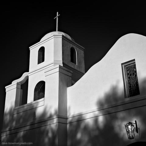 A b&w photograph of The Old Adobe Mission in Scottsdale, Arizona, showing its white walls illuminated by sunlight with the shadows of nearby trees cast across its façade, highlighting the texture and shape of the building against a dark, contrasting background.
