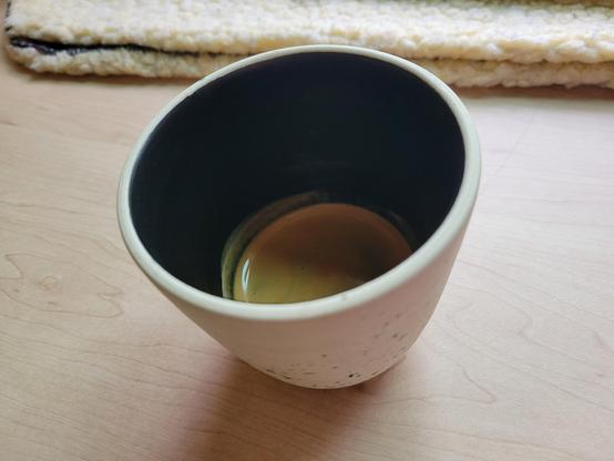 12-ounce, handle-less, off-white ceramic cup with a espresso shot, on a maple desk. The clay of the cup has many tiny flecks of black and blue. The interior of the cup has a dark-colored glaze.