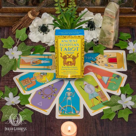A boxed deck of tarot cards stands amongst examples of the faces of some of the cards arrayed on a wood surface. The colorful imagery used is reminiscent of figures painted on Egyptian tomb walls. The deck's box is labeled "Ancient Egyptian Tarot Cards". Presented by Inked Goddess Creations.