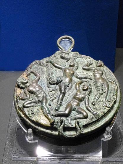 Etruscan bronze mirror case depicting the abduction of Ganymede by Zeus in the shape of a giant eagle. The eagle's talons hold on to a naked Ganymedes by the hips. Ganymedes reaches up, one arm holding on to the eagle's neck. Three figures are there to witness the abduction, two male and one female, all of them nude.